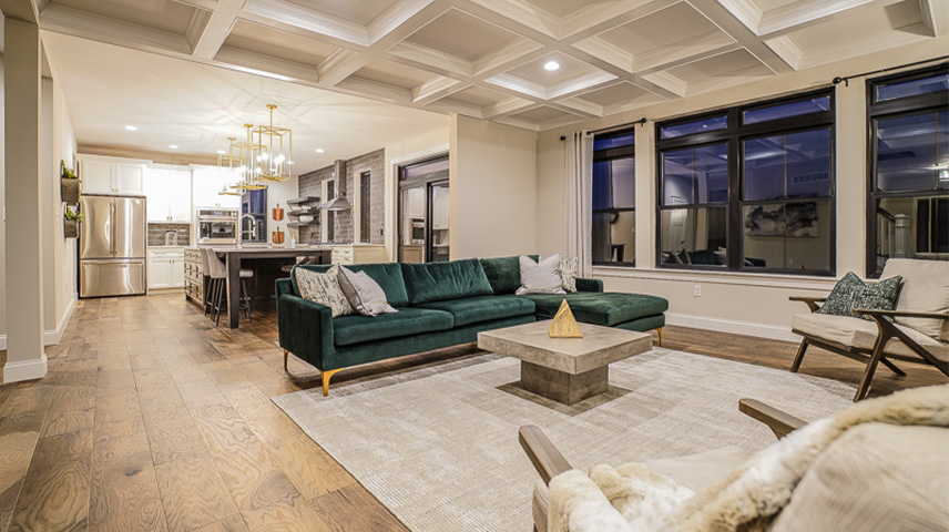 Modern open-concept living room with dark green sofa, concrete coffee table, and wood flooring; kitchen with stainless steel appliances and pendant lights visible in the background.