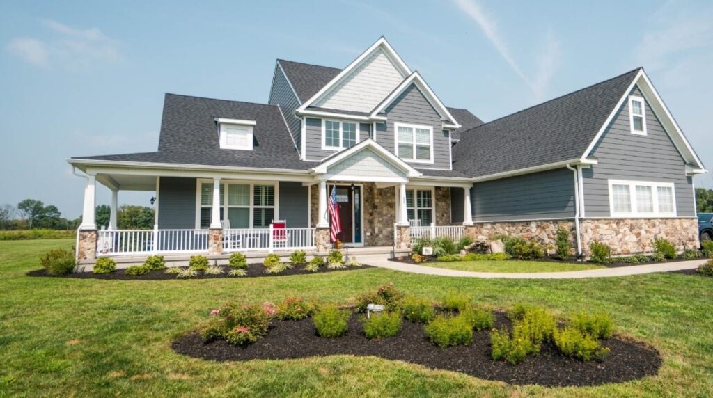 A large, modern two-story home with gray siding, stone accents, and a dark roof. The house features a wrap-around front porch with white railings, a well-manicured front yard, and a circular flower bed with shrubs. The home has multiple gables and a prominent front entryway with a red door and an American flag.