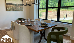 A modern dining room with a wooden table, six chairs (four beige, two green), set with plates and glasses, large windows overlooking a grassy area, and a landscape painting on the wall.