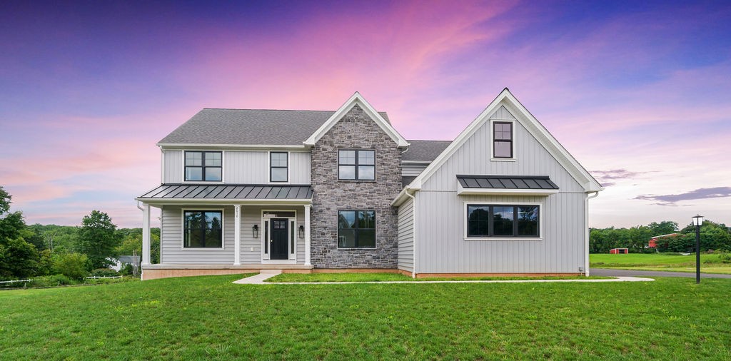 A modern two-story house with white siding, stone accents, and black trim sits on a green lawn under a vibrant pink and purple sunset sky. Trees and open fields surround the home.