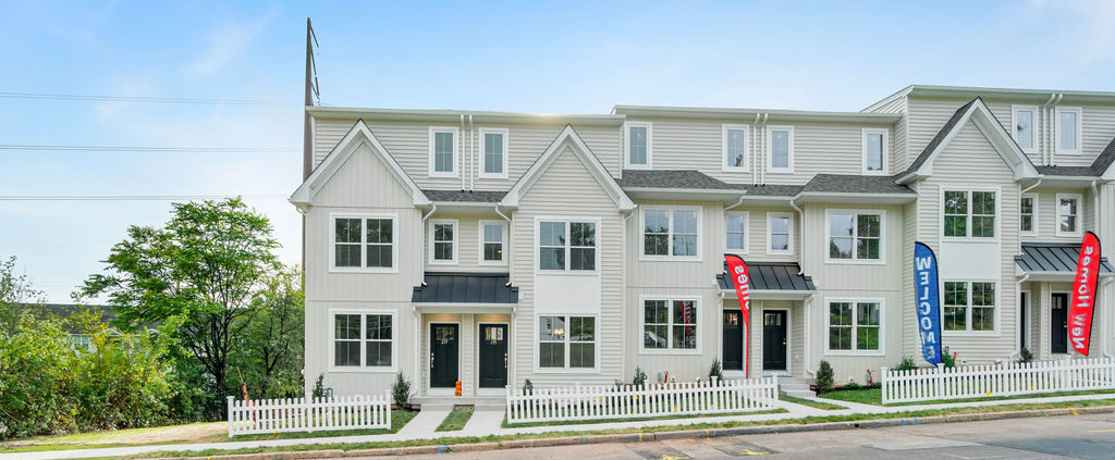 Row of modern white townhouses with black roofs, picket fences, and bright Welcome and Now Open flags by the entrances, set along a paved street with yellow lines on a clear, sunny day.