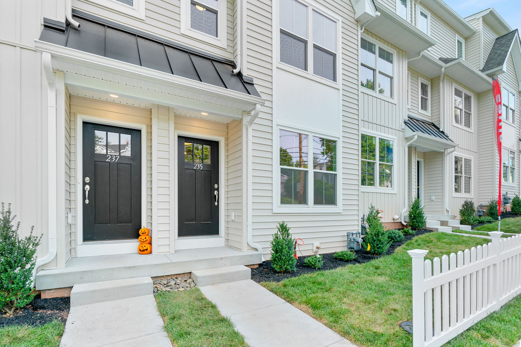Two black front doors on a beige townhouse with pumpkins stacked outside one door. The walkway is lined with green grass, small shrubs, and a white picket fence.
