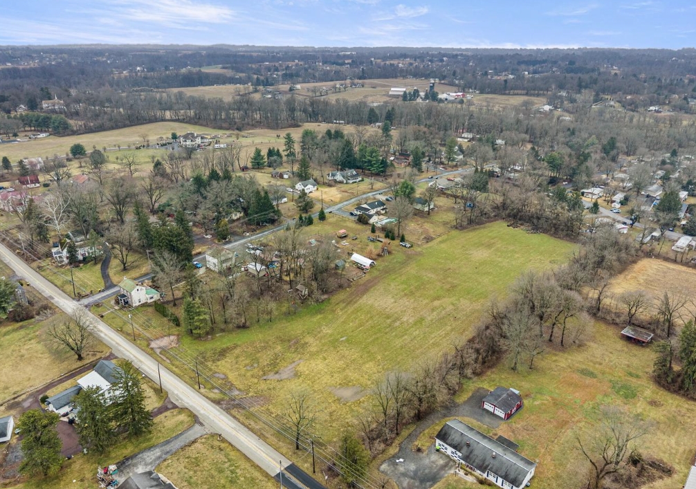 Aerial view of a rural area with scattered houses, open fields, leafless trees, and intersecting roads under a cloudy sky. The landscape is mostly brown and green, indicating early spring or late fall.