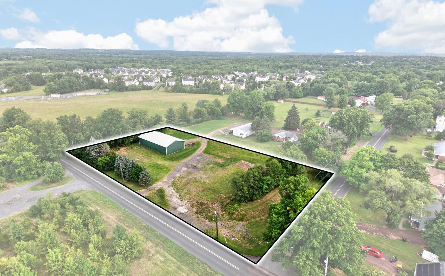 Aerial view of a rectangular, fenced lot with a green building and trees, surrounded by open fields and residential homes on a partly cloudy day.