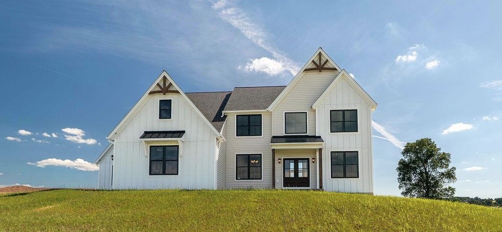 Modern white two-story house with black window frames and a gray roof, situated on a grassy hill under a blue sky with scattered clouds. A single tree stands to the right of the house.