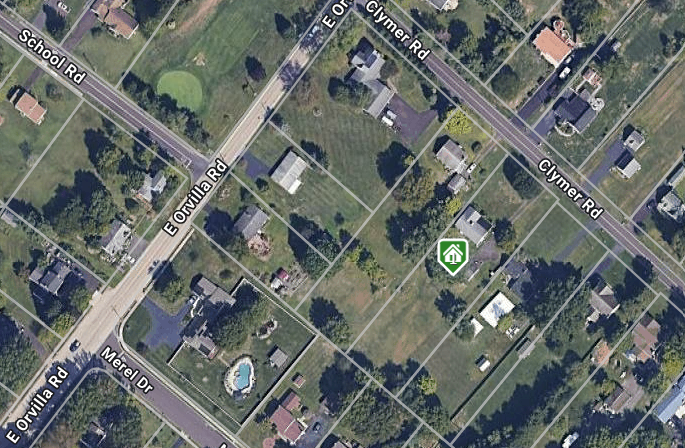 Aerial view of a suburban neighborhood with houses, green lawns, a swimming pool, and intersecting roads. A green home icon marks one property along Clymer Road.