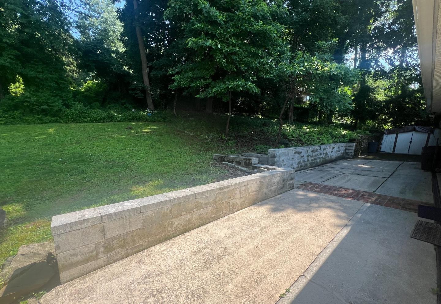 A concrete patio with a low retaining wall leads to a grassy yard bordered by trees. The scene is sunny, with shade from surrounding greenery, and a small shed is visible in the background.